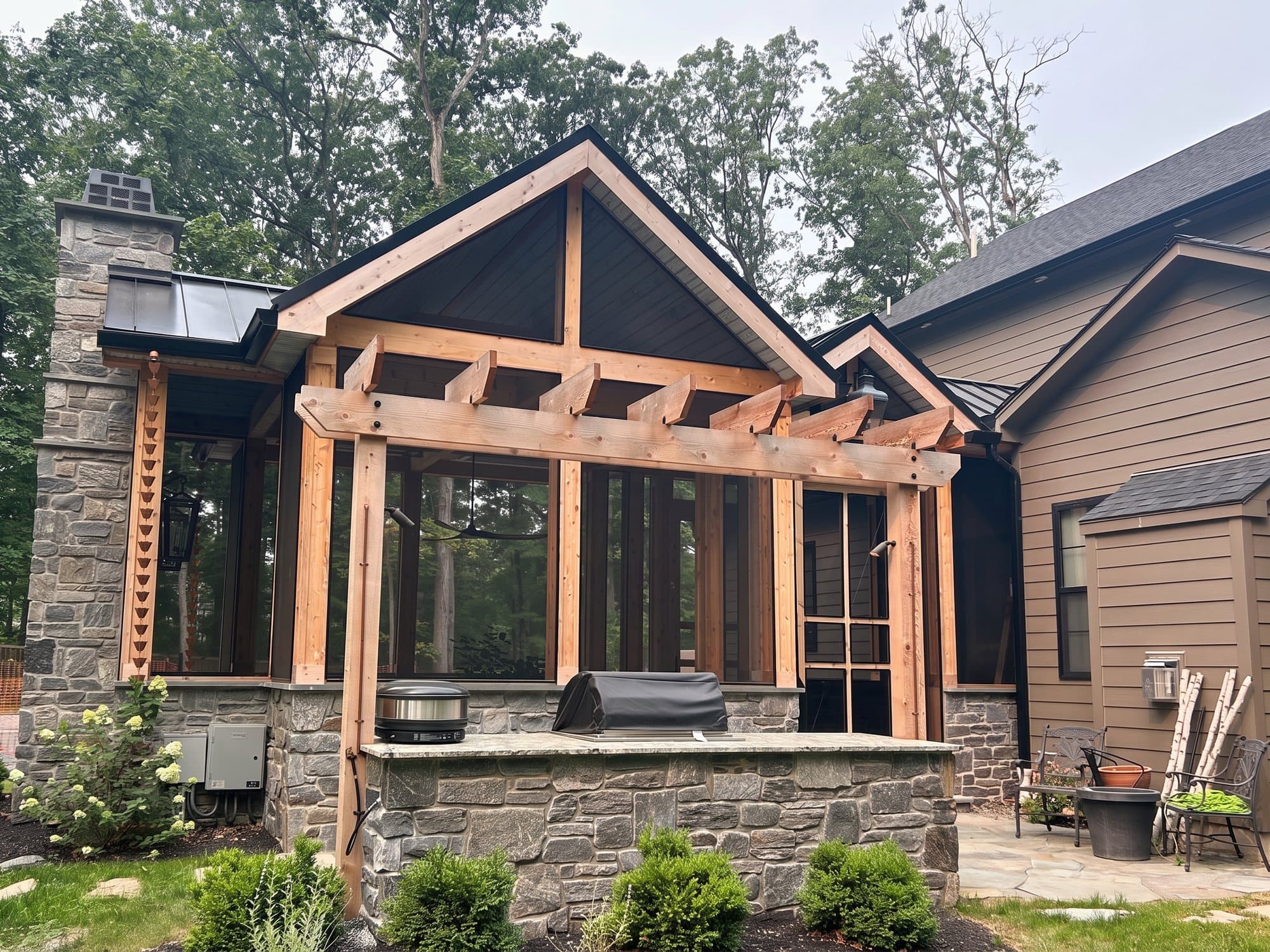 Timber pergola over a stone outdoor kitchen with a grill, beside a screened porch